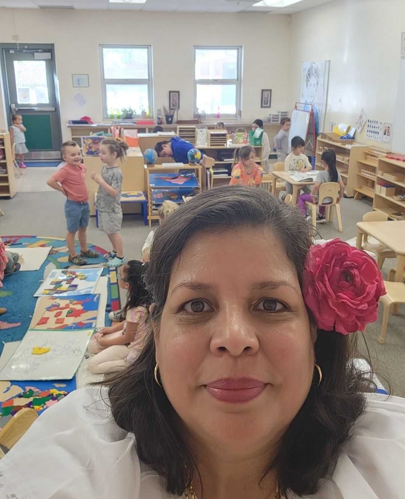 A woman with dark hair and a bright pink flower on the side of her hair is in the center of the image with her classroom of young children in the background. There's a colorful rug on the floor and some windows in the back.