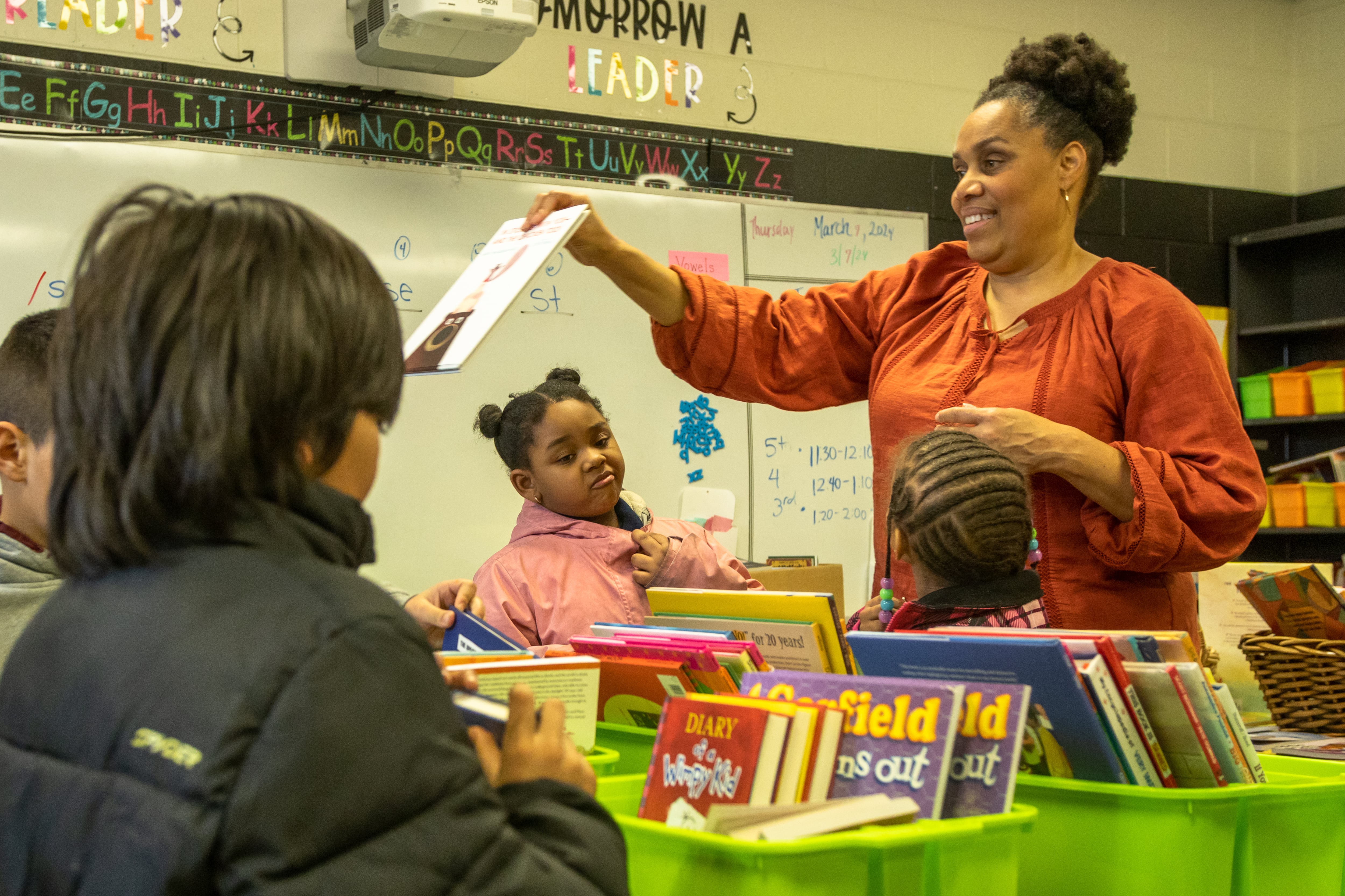 A woman stands holding a book, while several students look on. Baskets full of books sit on their classroom desks.